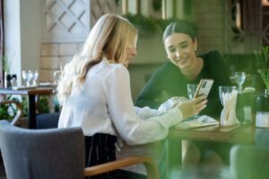 Två kvinnor vid restaurangbord, den ena håller upp en smartphone